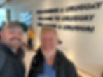 Two men smiling at a "Welcome to Uruguay" sign inside an airport. A woman in a yellow sweater stands in the background near a window.