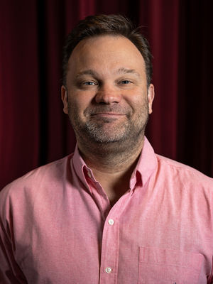 Komiker in einem pinken Hemd, im Hintergrund roter Vorhang. Er lacht.