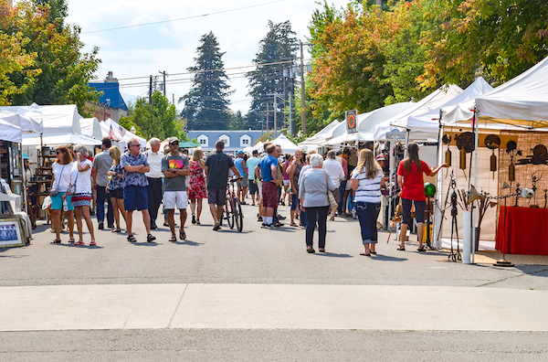 Outdoor market with many people browsing white tents. Trees and blue sky in background. Relaxed atmosphere on sunny day.