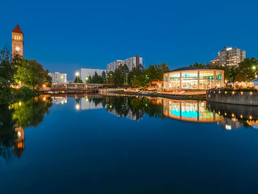 Clock tower and carousel by a calm river at dusk. Buildings and trees reflect in water. Evening lights create a serene, vibrant mood.