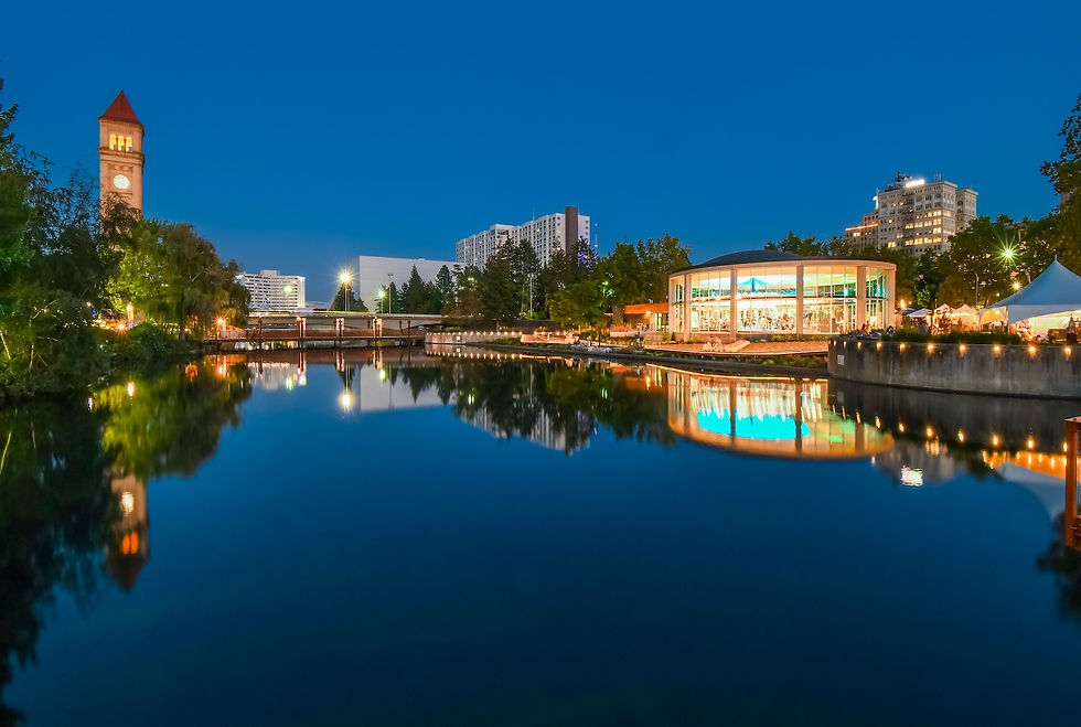 Clock tower and carousel by a calm river at dusk. Buildings and trees reflect in water. Evening lights create a serene, vibrant mood.
