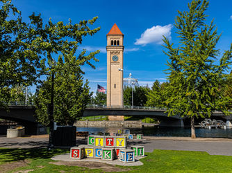 Clock tower in a park with "CITY SPOKANE" on colorful blocks in foreground. Blue sky, trees, bridge, and water create a tranquil setting.