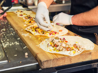 Person in gloves preparing wraps with chicken, vegetables, and cheese on a wooden board in a kitchen. Warm tones, casual atmosphere.