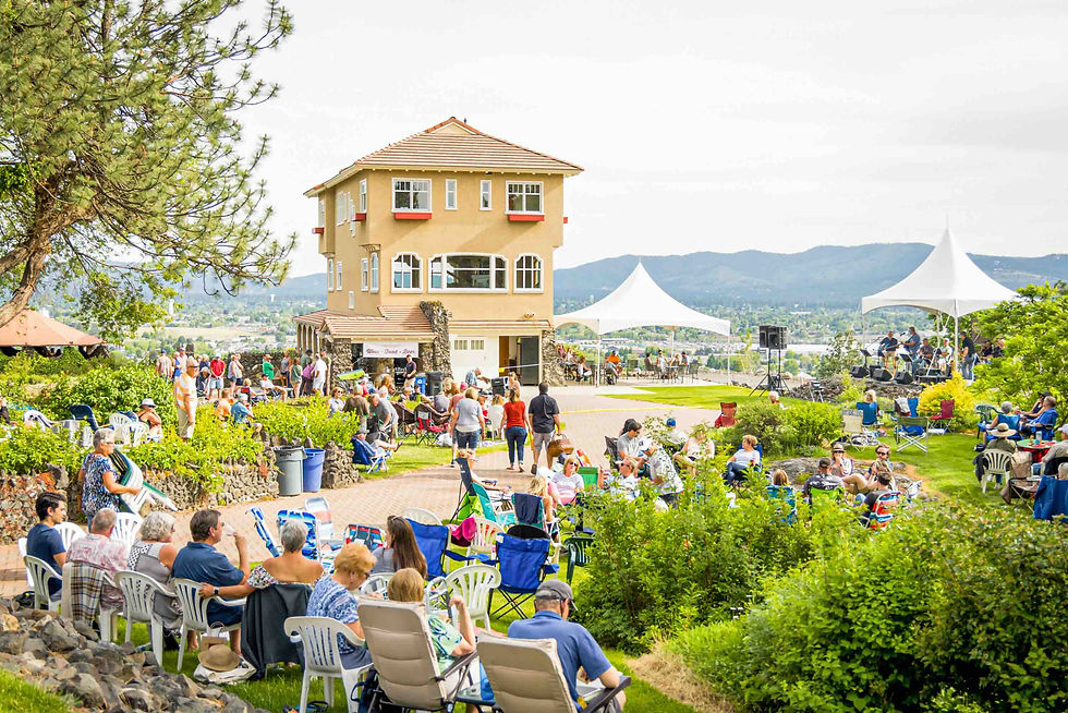 Outdoor event with people seated on chairs and blankets, gathering around a tan house. White tents in the background, scenic mountain view.