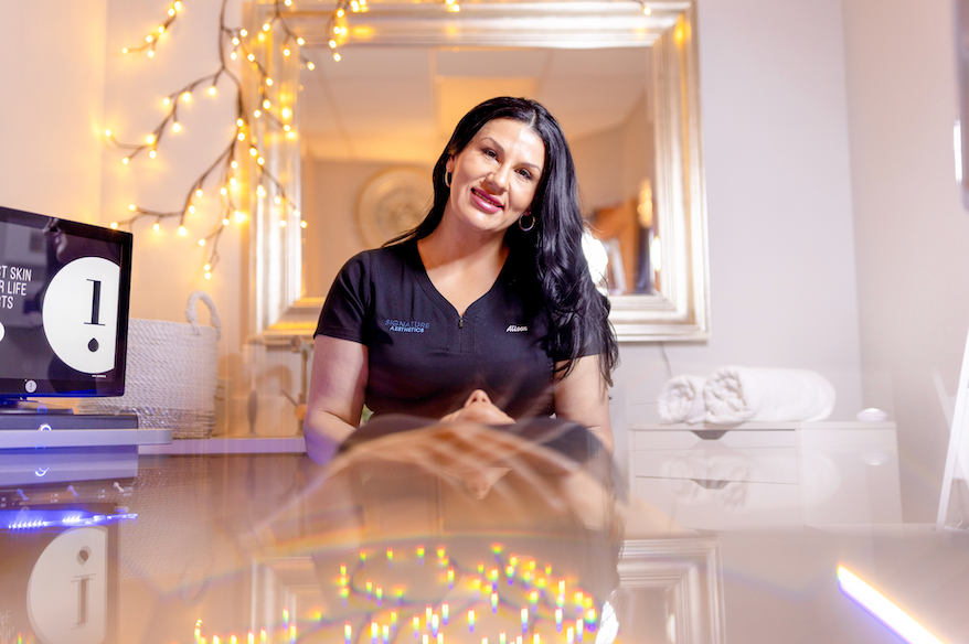 Smiling woman in a salon setting with string lights, mirror, and a screen showing text. Cozy ambiance with reflected colorful lights.