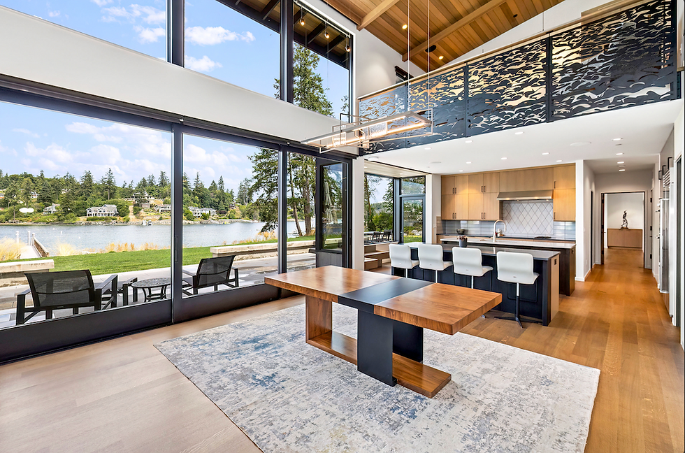 Modern interior with wooden table, kitchen island, and high ceilings. Large windows showcase a lake view surrounded by trees. Bright and airy.