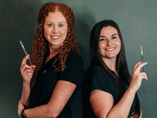 Two women with smiling expressions stand back to back, holding syringes. They wear black attire against a muted green background.