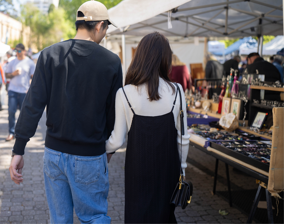 A couple walks at an outdoor market, browsing a stall with jewelry and art.