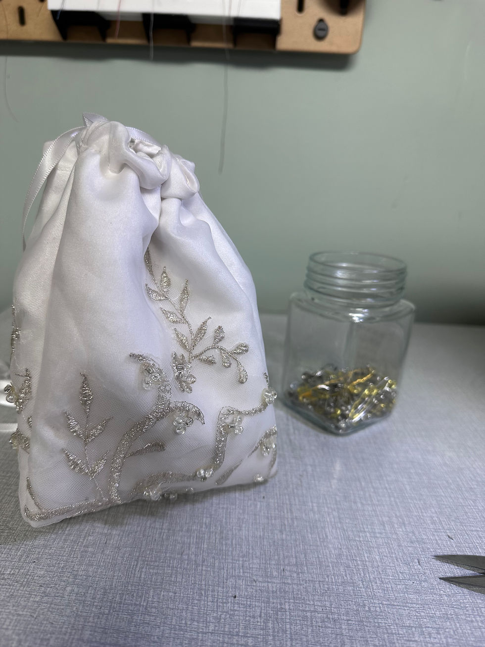 Embroidered white drawstring bag on table, next to a jar filled with metal clips. Soft lighting, neutral background, sewing theme.