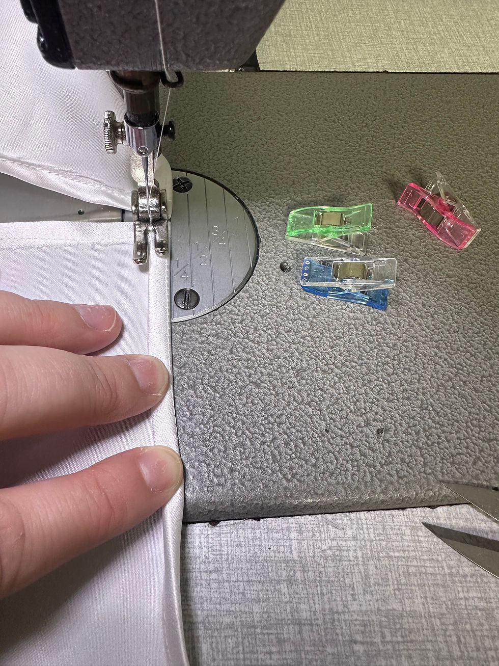 A sewing machine stitching white fabric, with a hand guiding it. Colorful clips and scissors are nearby on the textured gray table.