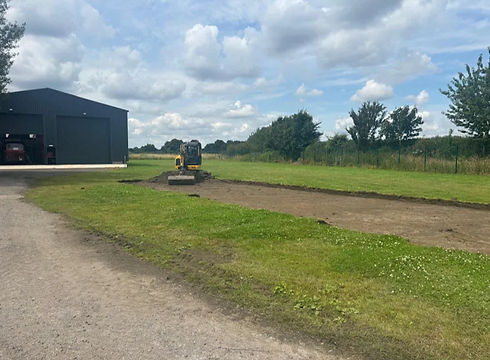 Mini excavator clearing topsoil for a new pathway next to an industrial barn.