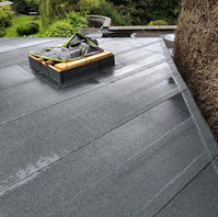 A flat roof scene showing a skylight surrounded by roofing material, with some greenery in the background