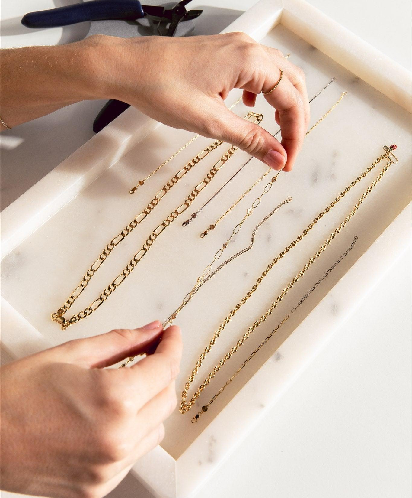 Hands examining delicate gold and silver chains arranged on a white marble jewelry tray beside a pair of pliers.