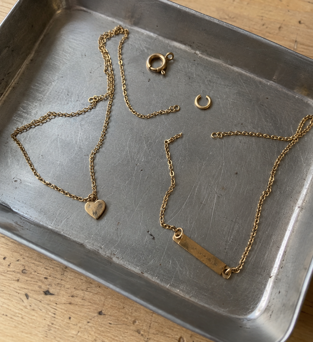 Broken gold chains, a heart pendant, clasp, and jump ring arranged in a metal jewelry repair tray.
