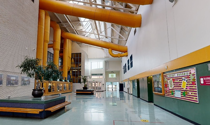 St. Patrick’s High School atrium with freshly painted white walls, yellow overhead structural beams, green accent wall, bulletin boards, seating area, and polished school flooring.