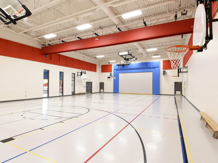Riverside South Elementary School gymnasium with freshly painted red and blue accent walls, white steel deck ceiling, basketball hoops, court lines, and bright overhead lighting.