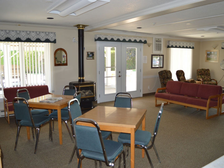 Centre J. Urgel Forget resident lounge with freshly painted light walls, tables, chairs, sofa seating, wood stove, glass doors, and bright ceiling lights.