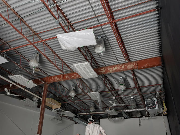 Warehouse ceiling preparation at Wellspring Studios in Nepean, Ontario, showing protected lighting fixtures, fans, mechanical components, exposed steel beams, roof deck and a painter working from lift equipment before commercial spray painting.