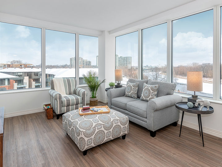 Greystone Village Retirement suite with freshly painted white walls, large corner windows, grey sofa, lounge chair, ottoman, side tables, and wood flooring.