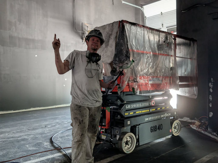 Commercial warehouse painting in progress at Wellspring Studios in Nepean, Ontario, showing a painter beside lift equipment, protected work areas, sprayed wall finishes, exposed ceiling structure and high interior walls.