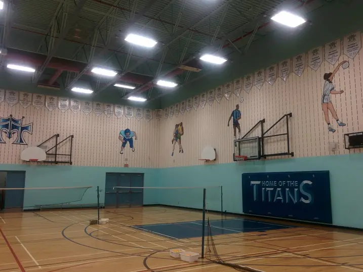 Mother Theresa High School gymnasium with freshly painted blue and grey walls, painted steel deck ceiling, basketball hoops, court lines, school banners, and Titans wall graphics.