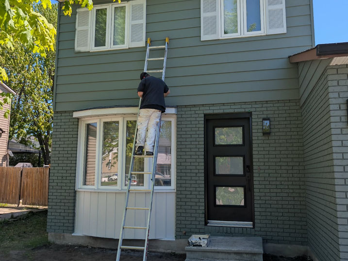Painter working on a two-storey house exterior with green painted siding and brick, white shutters and trim, black front door, ladder, and paint supplies.