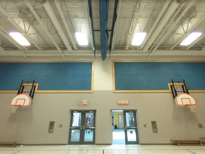 École Élémentaire Catholique Jean-Robert-Gauthier gymnasium with freshly painted exposed ceiling, blue wall panels, basketball hoops, painted doors, and bright overhead lighting.