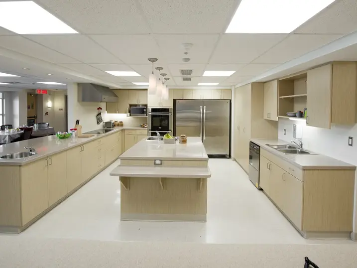 Health centre commercial kitchen with freshly painted walls, light wood cabinets, stainless steel refrigerators, prep counters, and bright ceiling lights.