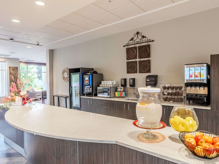 Wildpine Retirement Residence bistro area with freshly painted light walls, curved service counter, beverage station, fruit display, large windows, and bright ceiling lights.