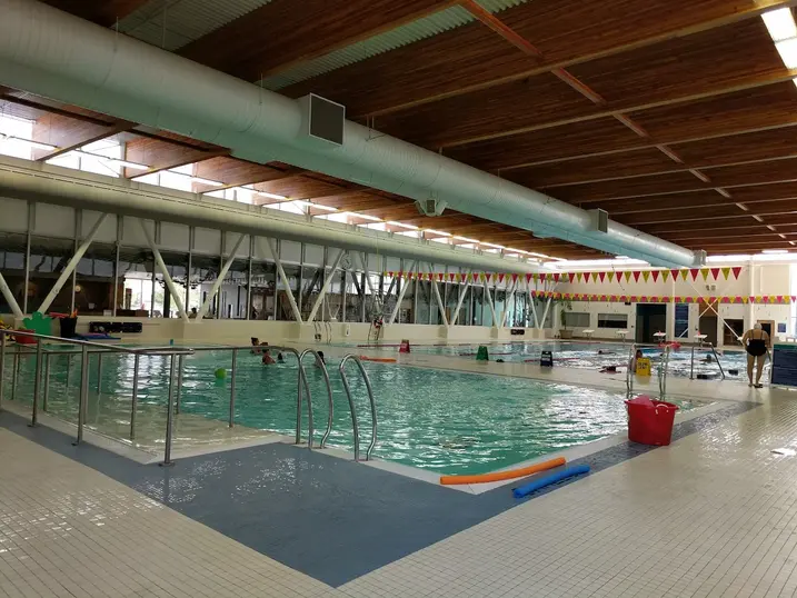 Francois Dupuis Recreation Centre indoor pool with stained wood ceiling, painted structural beams, large ductwork, swimming lanes, tile deck, and bright natural light.