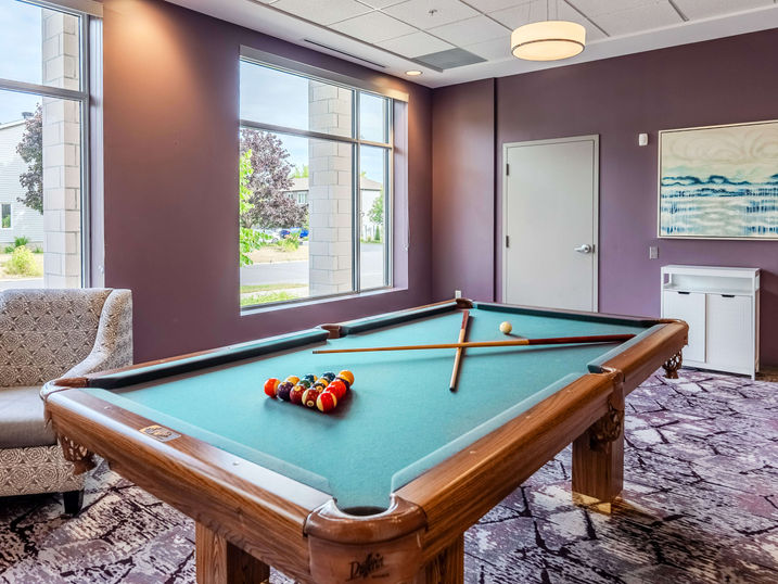 Wildpine Retirement Residence billiards room with freshly painted warm accent walls, pool table, large windows, armchair, patterned carpet, and bright ceiling lighting.