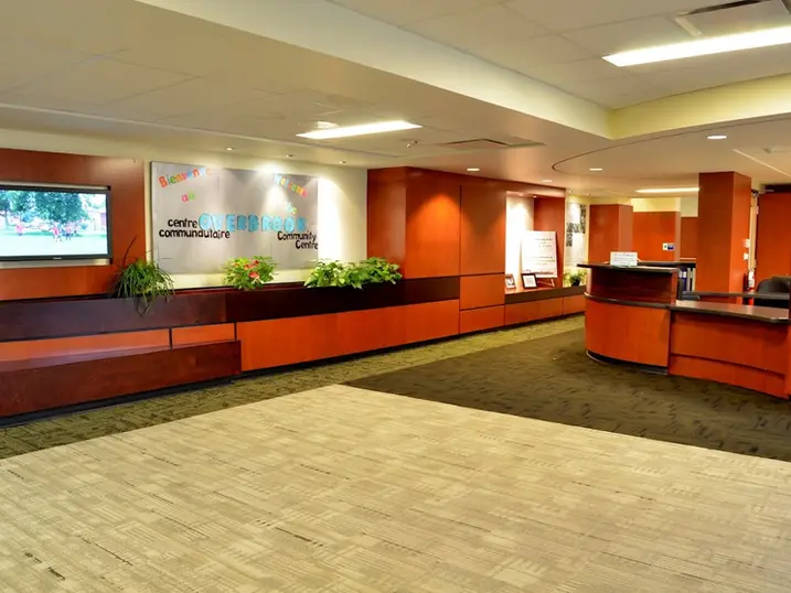 Overbrook Community Centre lobby with freshly painted warm accent walls, reception desk, seating area, community signage, plants, carpet flooring, and bright ceiling lights.