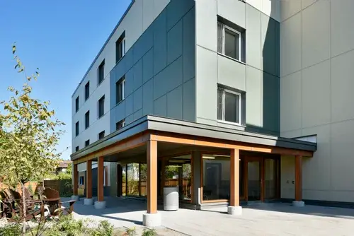 Apartment building entrance with stained wood posts and canopy structure, glass doors, grey exterior panels, and landscaped walkway.