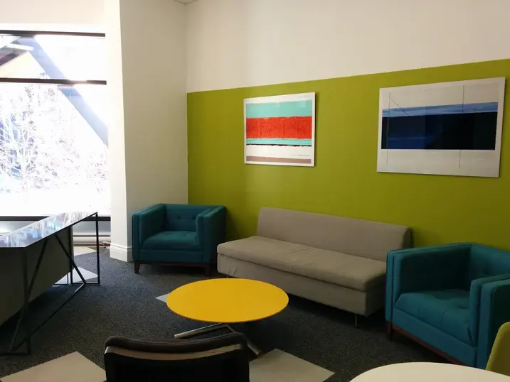 111 Cooper Street student lounge with freshly painted green accent wall, teal chairs, grey sofa, yellow table, large window, artwork, and carpet flooring.