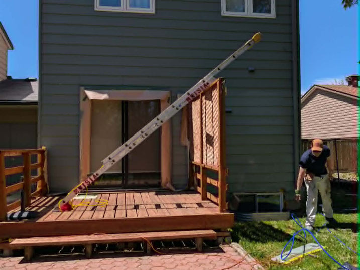 Painter working on the rear exterior of a two-storey house with green painted siding, ladder, wood deck, covered patio doors, and painting supplies.
