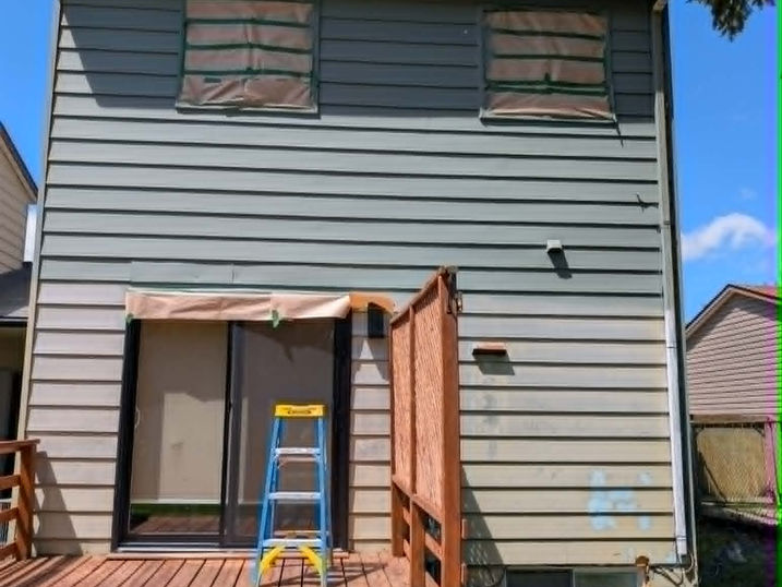 Rear view of a two-storey house with freshly painted green siding, covered windows, wood deck, ladder, and exterior painting in progress.