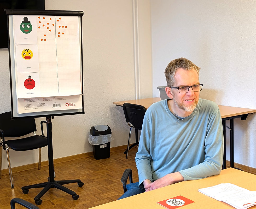 Mann sitzt lächelnd am Tisch, Flipchart mit bunten Gesichtern und Punkten im Hintergrund. Tisch mit Papieren und roter Karte.