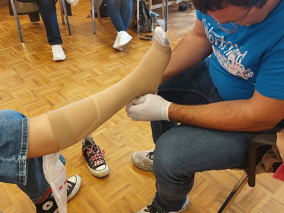Mann im blauen Shirt zieht einen Stützstrumpf über einen Unterschenkel, andere sitzen im Hintergrund.
