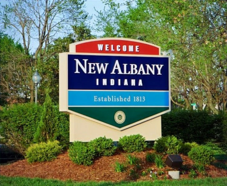 Welcome sign for New Albany, Indiana, reading "Established 1813," set in a lush green landscape with trees, under a clear sky.