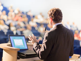 A speaker in a suit presents at a podium with a laptop to an audience in a lecture hall. The setting is bright and formal.