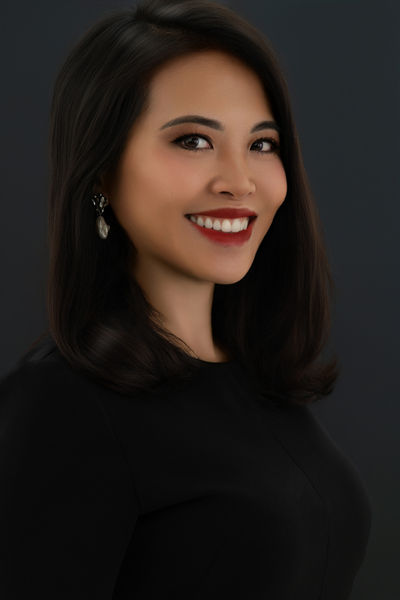 A headshot of a smiling woman with dark, shoulder-length hair, looking directly at the viewer. She has bright eyes, prominent eyeliner, and vibrant red lipstick. She is wearing a black top and a dangling earring on her left ear. The background is a solid dark gray.