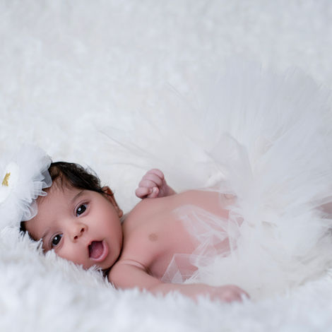 A newborn baby lying on a white fluffy blanket, wearing a white tutu and a white floral headband, with wide eyes and an open mouth, looking at the viewer.