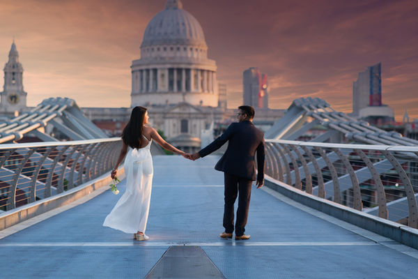 A couple holds hands and walks away from the viewer on the Millennium Bridge in London, with St. Paul's Cathedral dominating the background under a sunset sky. The woman on the left wears a flowing white backless dress and holds a white rose in her other hand. The man on the right wears a dark suit. The bridge's distinctive steel structure is visible on either side, and other city buildings can be seen in the distance, partially silhouetted by the warm glow of the setting sun.