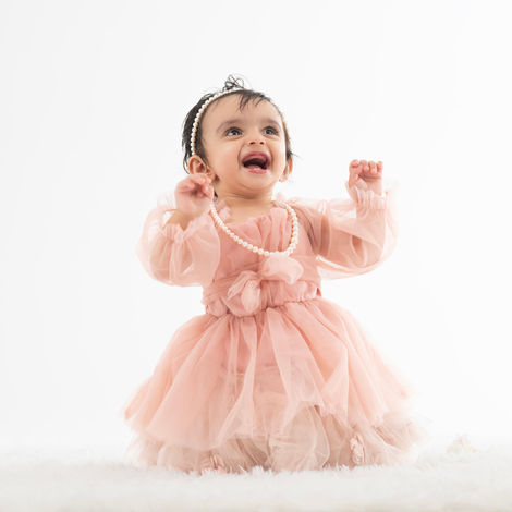 A happy baby girl with dark hair, wearing a light pink tulle dress and a pearl necklace, sits on a white furry surface and looks up with an open-mouthed smile, with hands raised slightly.