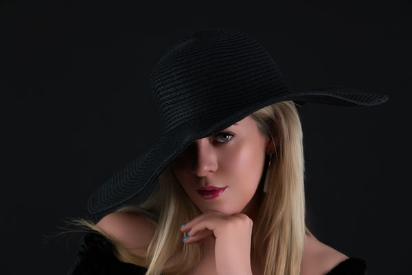 A headshot of a woman with long blonde hair, partially obscured by a wide-brimmed black hat. Her right eye is visible, looking directly at the viewer, while her left eye is cast in shadow by the hat. She has bold red lipstick and a thoughtful expression, with her left hand resting on her chin. She appears to be wearing an off-the-shoulder black top. The background is solid black.