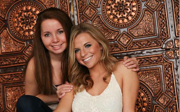 A young, white, brunette woman, Lexi Lamb, smiles and holds the shoulders of a young, white, blond smiling woman, Ashley Lamb.