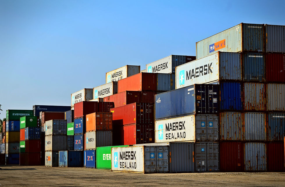 Stacked shipping containers in various colors with brand logos, under a clear blue sky. Labels like Maersk and Evergreen visible.