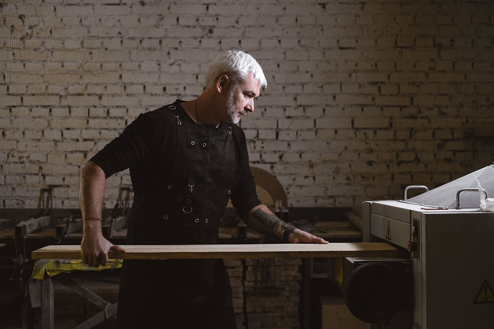 Man with white hair working on wood in a dimly lit workshop. He wears a black apron. Brick wall background. Concentrated mood.