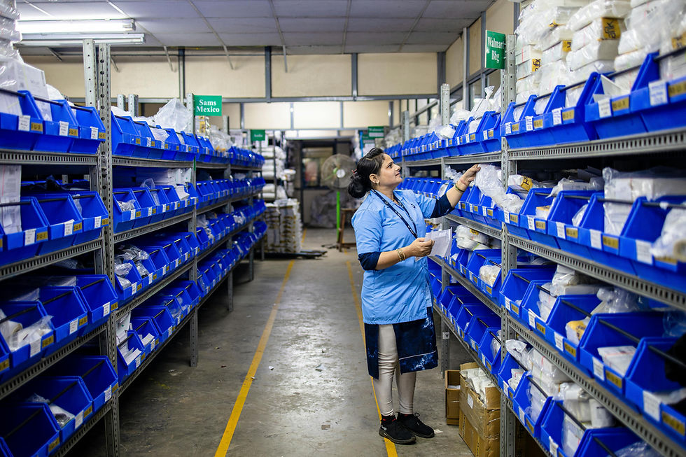 A woman in a blue uniform organizes items on shelves with blue bins in a warehouse. Signs read "Sams Mexico" and "Walmart Bkg."