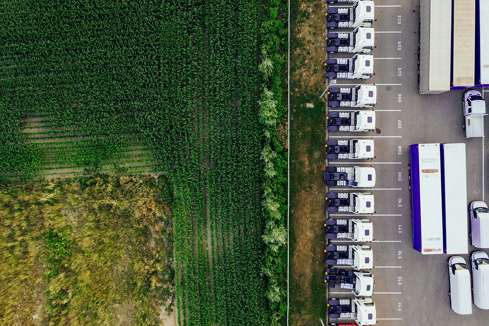 Aerial view of green crop fields beside a truck yard with parked white semis marked E03–E14 and purple trailers.